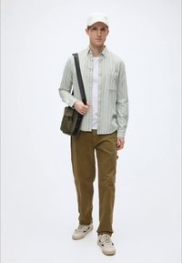 Φωτεινό πράσινο ριγέ πουκάμισο πάνω από λευκό T-shirt, χακί παντελόνια και μπεζ αθλητικά παπούτσια. Κρατάει μια πράσινη τσάντα ώμου. Απλός, καθημερινός σχεδιασμός.