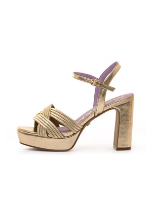 Gouden hoge hak sandaal met plateau, gekruiste gestructureerde bandjes, enkelgesp en lavendelkleurige binnenzool, weergegeven tegen een witte achtergrond.