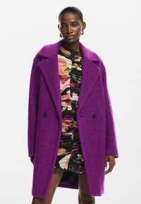 Abrigo púrpura con textura, solapa en pico y dos botones negros, superpuesto a un vestido con estampado floral que presenta detalles con volantes.