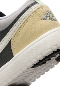 Κοντινό πλάνο της φτέρνας αθλητικού παπουτσιού με επικάλυψη από μπεζ σουέτ, μαύρες και λευκές δερμάτινες περιοχές, λευκή ενδιάμεση σόλα και σκούρο ραμμένο λογότυπο φτερού.