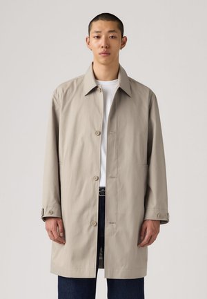 Manteau beige léger avec un col classique, des boutons sur le devant et deux poches latérales, porté sur un t-shirt blanc et un jean foncé.
