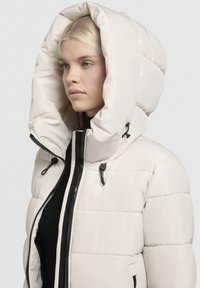Veste bouffante couleur crème avec une grande capuche, dotée de zips et de lacets noirs, et d'une texture matelassée pour l'isolation.