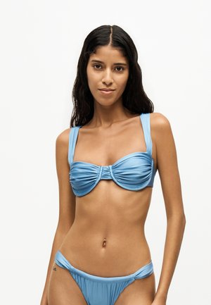 Femme aux longs cheveux foncés portant un haut et un bas de bikini bleu clair froncés, debout devant un fond blanc uni.