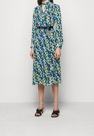 Robe à imprimé floral en bleu, vert et blanc, avec un col, des manches longues et une taille ceinturée. Portée avec des sandales à talons noires.