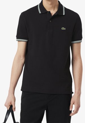 Homme portant un polo noir à manches courtes avec col et poignets à rayures blanches, tenant une sangle noire dans la main gauche sur fond blanc.