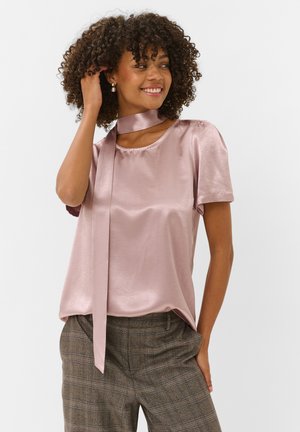 Femme aux cheveux bouclés sourit, portant un chemisier rose clair brillant avec une écharpe au cou et un pantalon gris à carreaux, debout devant un fond uni.