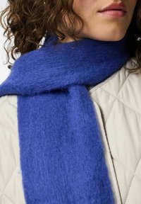 Persona con cabello rizado que lleva una bufanda de lana azul brillante sobre una chaqueta acolchada beige, mostrando parte del cuello, labios y mentón.