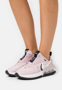Las zapatillas Nike de color rosa claro presentan una parte superior de malla transpirable, acentos negros y una unidad de aire transparente en la suela. Ubicadas sobre un fondo neutro.