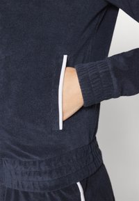 Sweat-shirt bleu marine avec un tissu texturé, comprenant une poche à passepoil blanc et des poignets côtelés pour plus de détail et de structure.