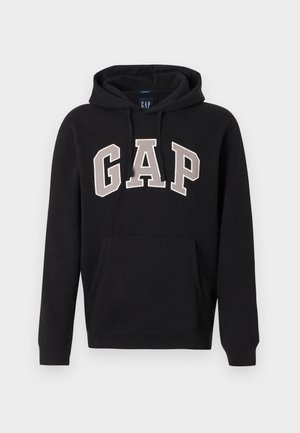 Svart hettegenser med frontlomme og stor "GAP"-logo i beige, konturerte bokstaver over brystet.