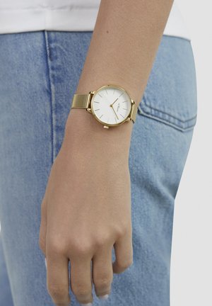 Goldfarbenes Mesh-Armband-Uhr mit rundem weißem Zifferblatt, dünnen Markierungen und minimalistischem Design, am Handgelenk neben hellblauem Denim befestigt.