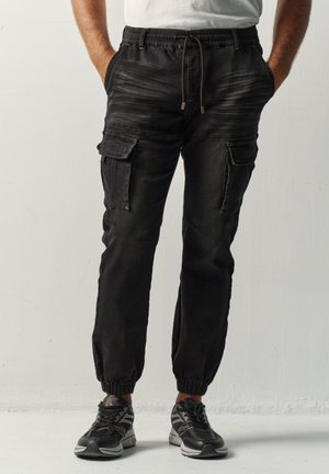 Hombre con pantalones jogger cargo negros con puños elásticos y cordón, combinado con zapatillas negras y blancas, de pie con las manos en los bolsillos.