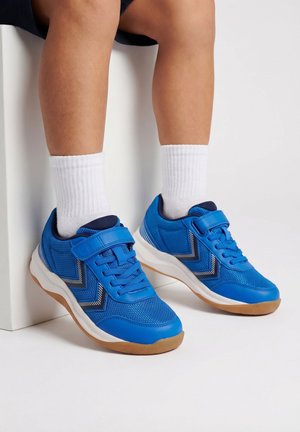 Kinderbeine mit weißen Socken und leuchtend blauen Sneakern mit Klettverschlüssen und weißen Gummisohlen, sitzend vor weißem Hintergrund.