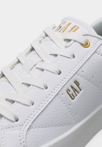 Baskets en cuir blanc avec lacets blancs, œillets dorés et logo "GAP" doré sur la languette et le côté.