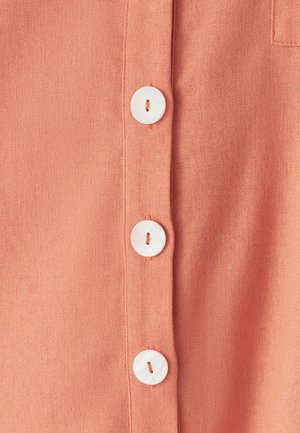 Koraļkrāsas kokvilnas krekls ar baltām pogām, fakturētu audumu un gludu apdari ar pogu ailes detaļu priekšpusē.