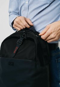 Czarna plecak wykonany z trwałego materiału o gładkiej fakturze, z uchwytem u góry, zamkiem błyskawicznym i czerwoną metką na przodzie.