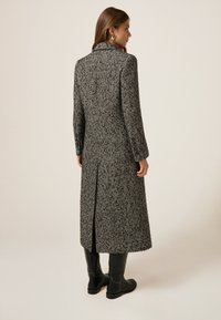 Cappotto lungo con motivo a spina di pesce bianco e nero, con colletto a punta, maniche lunghe e uno spacco sul retro, abbinato a stivali neri fino al ginocchio.