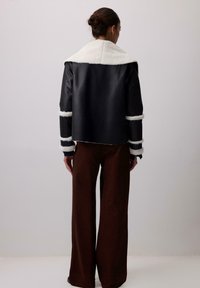 Cilvēks ar matiem sasietiem bunā, uzvilcis melnu vilnas jaku ar baltu apdari un brūnām platām biksēm, stāv pret vienkāršas fona.