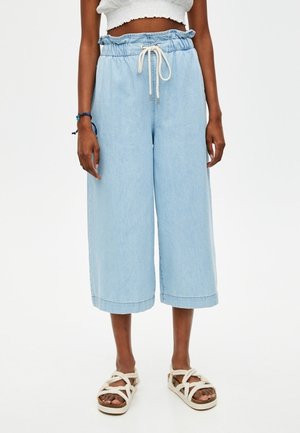 Žena nosící světle modré široké krátké džíny s volánkovým elastickým pasem a bílé sandály s provázky na bílém pozadí.