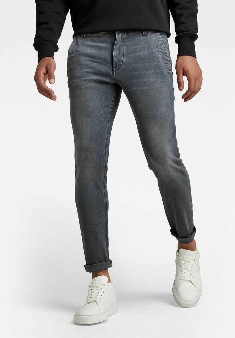 Jean en denim gris à coupe slim, avec des revers roulés et une finition délavée. Associé à des baskets blanches, soulignant le look décontracté général.