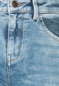 Jean en denim bleu clair en gros plan montrant le bouton, la boucle de ceinture, la poche avant et les surpiqûres orange contrastées.