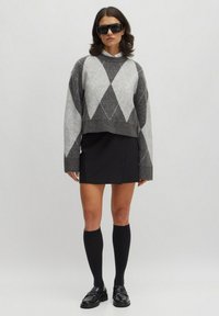 Gekürzter Pullover in Grau und Hellgrau mit einem Rautenmuster. Kombiniert mit einem schwarzen Minirock, kniehohen schwarzen Socken und Loafers.