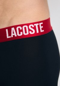 Crne bokserice s širokim crvenim elastičnim pojasom na kojem je bijelim slovima ispisano "LACOSTE". Glatka textura s prilagođenim dizajnom.