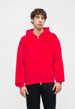 Junger Mann trägt einen leuchtend roten Kapuzenpullover mit Reißverschluss und kleinem schwarzem Logo auf der Brust, kombiniert mit schwarzen Hosen, stehend vor einem einfarbigen Hintergrund.