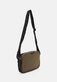 Rektangulär crossbody-väska i olivgrön och svart tyg. Har justerbar svart rem och silverdetaljer. Slät textur, enkel design.