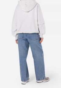 Světlé šedé mikina s kapucí a spadlými rameny, kombinovaná s volnými, vybledlými modrými džíny. Jemné švy na kapsách.