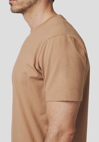 Brązowa bawełniana koszulka z krótkim rękawem, okrągłym dekoltem i gładką teksturą. Widok z boku pokazuje dopasowany krój i szwy na ramionach.