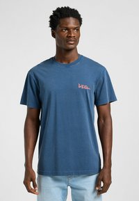 Sininen puuvillainen t-paita lyhyillä hihoilla, jossa on pieni vaaleanpunainen "Lee" -logo rinnassa ja normaali pyöreä pääntie.