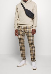 Beige hoodie, plaid brown and black fitted pants, and white sneakers with accents; black crossbody bag worn across the body.