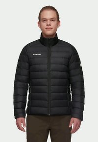 Giovane uomo che indossa una giacca trapuntata nera Mammut con zip frontale e tasche laterali con zip, in piedi contro uno sfondo neutro.