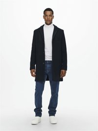 Manteau en laine bleu marine avec un col cranté et une fermeture à un seul bouton, associé à un col roulé blanc et un jean bleu ; des baskets blanches complètent la tenue.