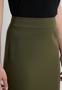 Olivegrüner Midirock mit glatter Textur und hohem Bund, ausgestattet mit dezenten Seitennähten. Kombiniert mit einem schwarzen Oberteil mit Glitzer.