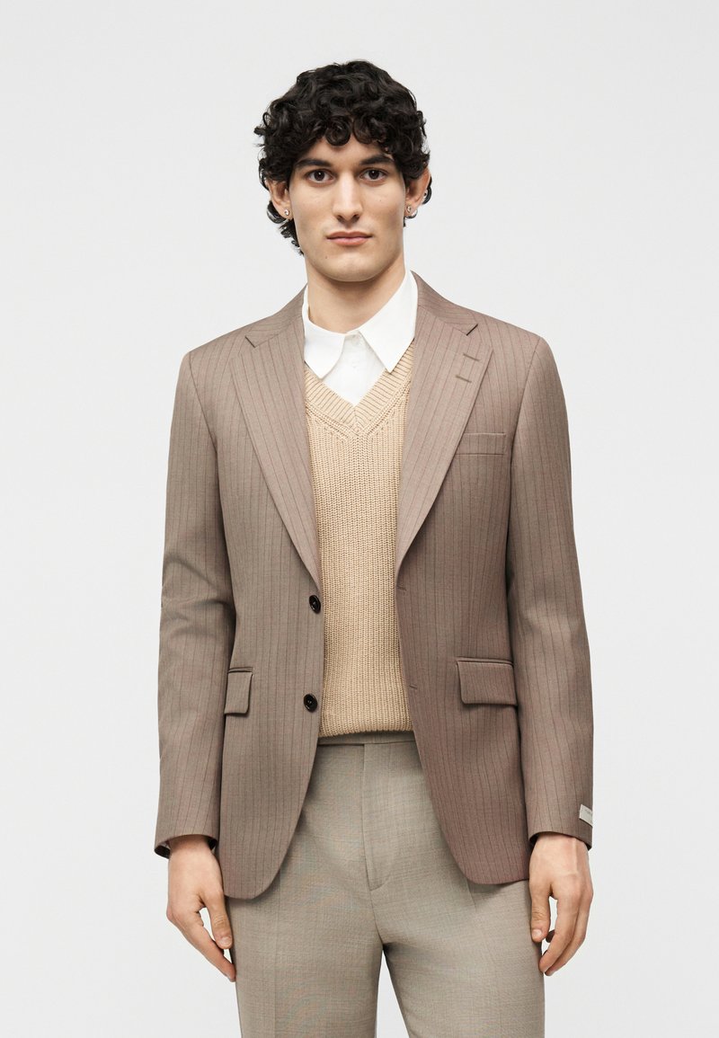 Νεαρός άνδρας που φοράει μπεζ σακάκι με ρίγες, μπεζ πλεκτό γιλέκο, λευκό πουκάμισο με γιακά και ανοιχτό μπεζ παντελόνι, στέκεται μπροστά σε απλό φόντο.