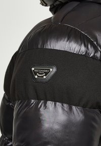 Sort puffervest med en blanding af skinnende og matte teksturer. Indeholder et trekantet logoemblem med tekst på den øverste del af ryggen.