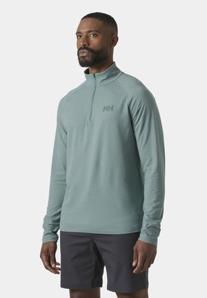 Mężczyzna z krótką brodą, ubrany w jasnooliwkową bluzę na suwak z długim rękawem i czarne spodenki, stojący na tle jednolitego, jasnego tła.