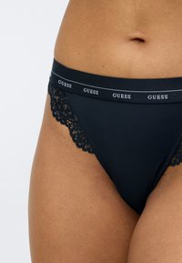 Zwarte stringonderbroek met een kanten randje, voorzien van een zachte tailleband versierd met het "GUESS"-logo in het wit. Overal een gladde textuur.
