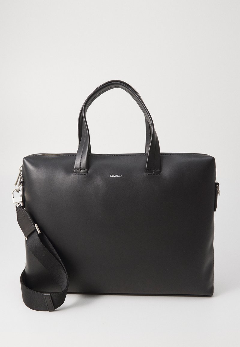 Črna usnjena torba za prenosni računalnik z dvema zgornjima ročajema, snemnim ramenskim trakom in zadrgo. Na sprednji strani je subtilen logotip.