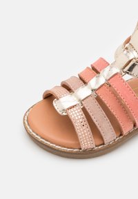 Sandaalit, joissa on nahkaremmit, metalliset kultaiset yksityiskohdat, vaaleanpunaiset ja beige-nahkaremmiset, sekä sileä ruskea pohja. Ompeleita pohjan ympäri.