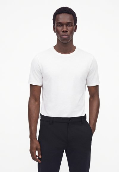 Homme portant un t-shirt blanc uni à manches courtes et un pantalon noir, debout devant un fond blanc uni.