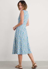 Ziedu raksta bez piedurknēm kleita gaiši zilos toņos ar dzelteniem un purpura akcentiem; ceļgala garums, plūstoši audumi, apvienota ar plakanām sandālēm.