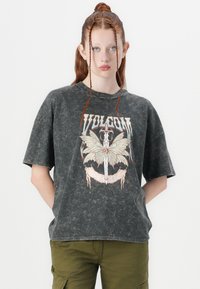 Junge Person mit langen, geflochtenen roten Haaren trägt ein verblasstes schwarzes Volcom-T-Shirt mit einem Schmetterlings- und Schwertmotiv, kombiniert mit olivgrünen Cargo-Hosen.