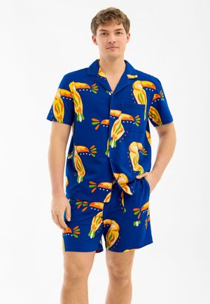 Nuori mies, jolla on sininen lyhythihainen napitettava paita ja siihen sopivat shortsit, joissa on värikäs tukaani-lintukuosi, seisoo käsi taskussa.