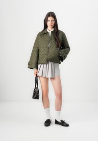 Donna che indossa una giacca trapuntata verde oliva, una mini gonna grigia a pieghe, calze bianche, mocassini neri e tiene in mano una borsa nera.
