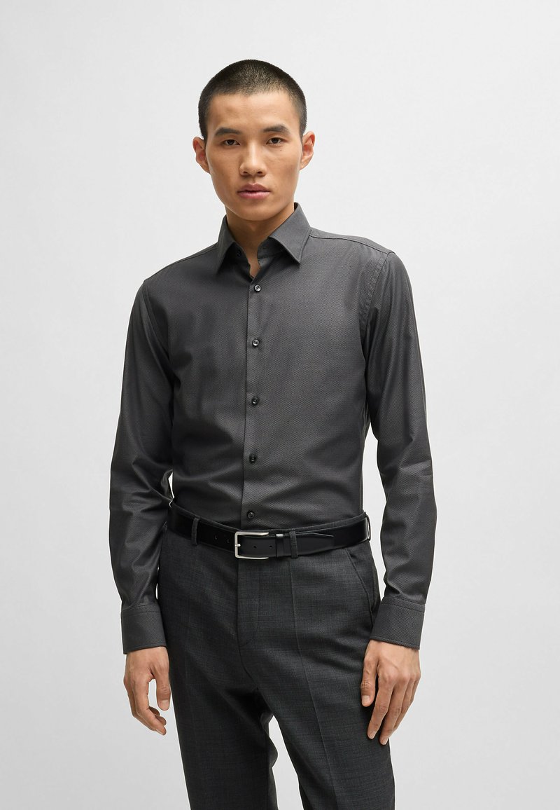 Chemise à boutons grise pour homme en tissu texturé, avec un col classique, des manches longues et des boutons noirs. Portée avec un pantalon sombre ajusté.