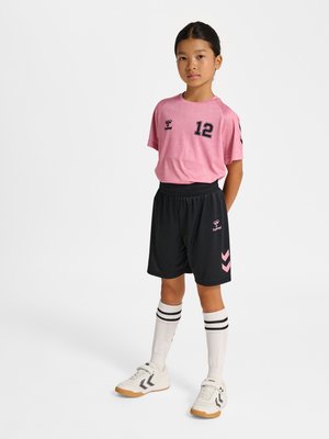 Pige stående med hænderne bag ryggen iført pink sportstrøje nummer 12, sorte shorts, hvide knæstrømper og hvide sneakers.