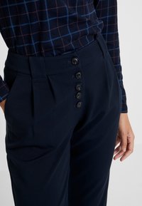 Granatowe spodnie o gładkiej teksturze, z wysokim stanem, czterema guzikami i bocznymi kieszeniami, zestawione z bluzką w granatową kratę.